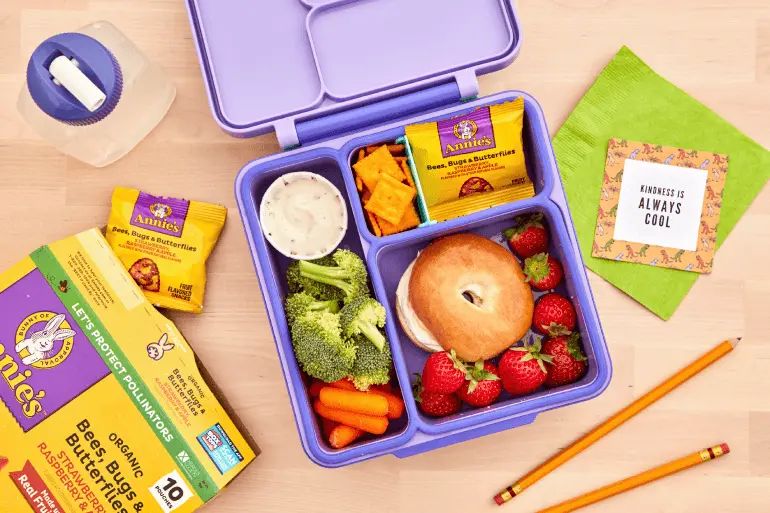 A lunchbox containing a bagel sandwich, strawberries, broccoli, carrots, a small container of dip, and a pack of Annie's snacks. Nearby are a water bottle, a box of Annie's Bees, Bugs & Butterflies snacks, a green napkin, a card with the text 'Kindness is always cool,' and two pencils.