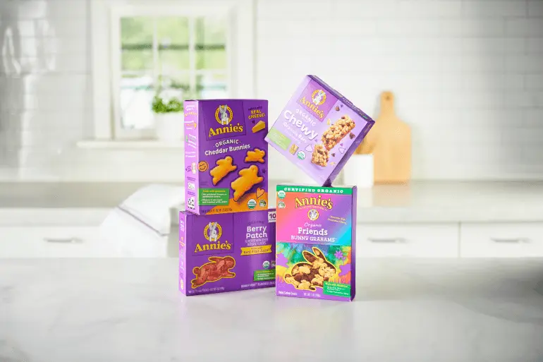 Four boxes of Annie's organic snacks are displayed on a kitchen counter. The boxes include Organic Cheddar Bunnies, Berry Patch, Chewy Granola Bars, and Organic Friends Bunny Grahams. A window and kitchen utensils are visible in the background.